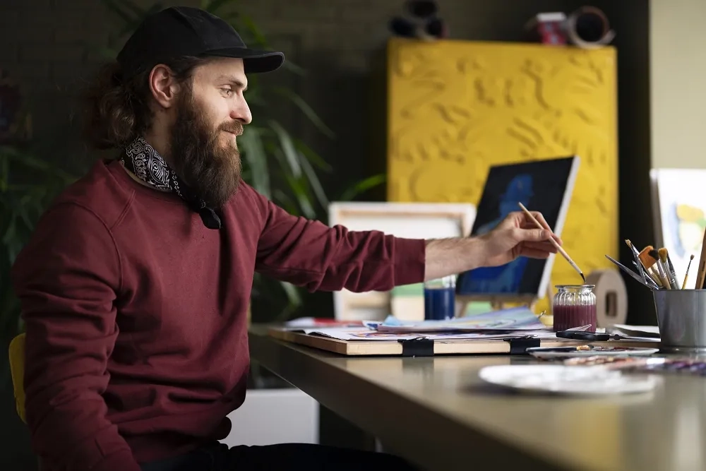 Artist working in a creative studio, painting at a desk with brushes and supplies, representing topics in creative artists management and support.