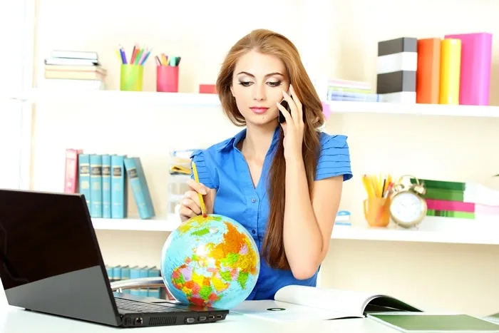 Woman studying with a laptop while talking on the phone and examining a globe, symbolizing research and evaluation of free online course opportunities.