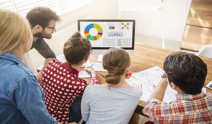 Team collaborating around a computer displaying charts and analytics, illustrating real-time teamwork and shared coding workflows using Google Colab.