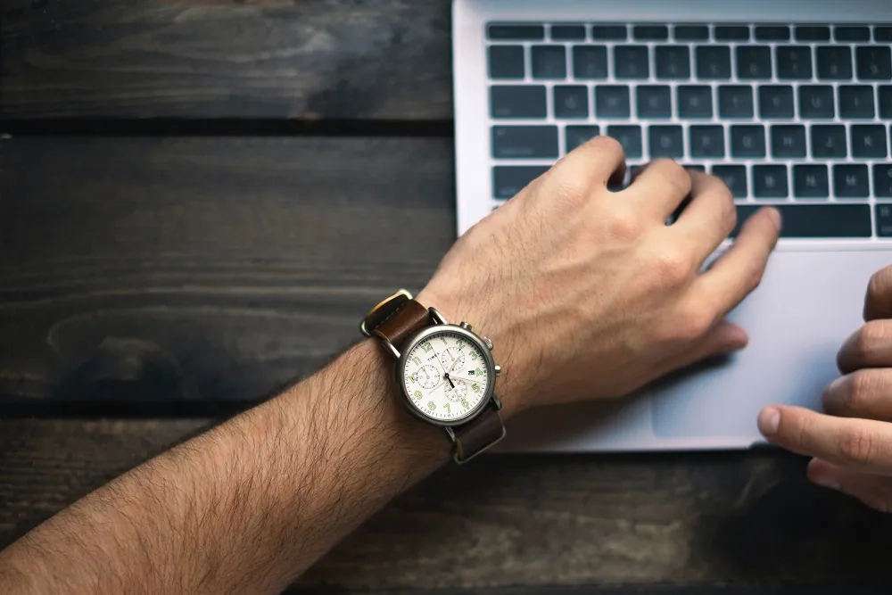 Close-up of a person checking the time on a wristwatch while working on a laptop, symbolizing productivity and the latest time management techniques.