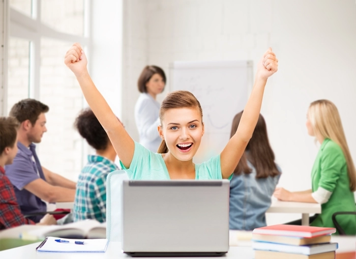 Happy student raising arms behind a laptop in a classroom, representing the accessibility and popularity of MOOCs.