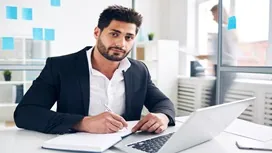 Professional office worker writing notes beside a laptop, representing administrative, secretarial, and PA skills at Level 5.
