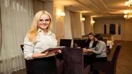 Hotel staff member holding a tablet in a modern dining area, representing professional training in hotel management and customer service.