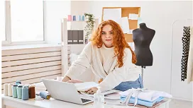 A fashion designer working at a desk with fabric, sketches, and a laptop, representing practical skills taught in a Level 4 fashion design diploma.