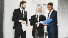 Three business professionals discussing documents in a hallway, symbolizing leadership, teamwork and organisational skills taught in management courses.