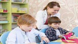 A nursery nurse supporting young children during creative activities in a classroom environment, reflecting advanced early-years skills taught in Level 7 nursery nurse diplomas.