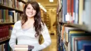 Smiling student holding books in a library aisle, representing learning, customer service skills, and core study themes in retail management.