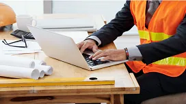 A construction professional in a safety vest working on a laptop beside blueprints, illustrating essential skills for Site Management QLS Level 3.