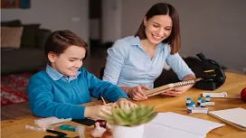 A teaching assistant helping a young student with schoolwork at a table, representing supportive learning and child development taught in Level 4 teaching assistant diplomas.