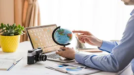 A person planning a trip using a globe, maps, and a laptop, representing key introductory concepts taught in Level 3 travel and tourism courses.